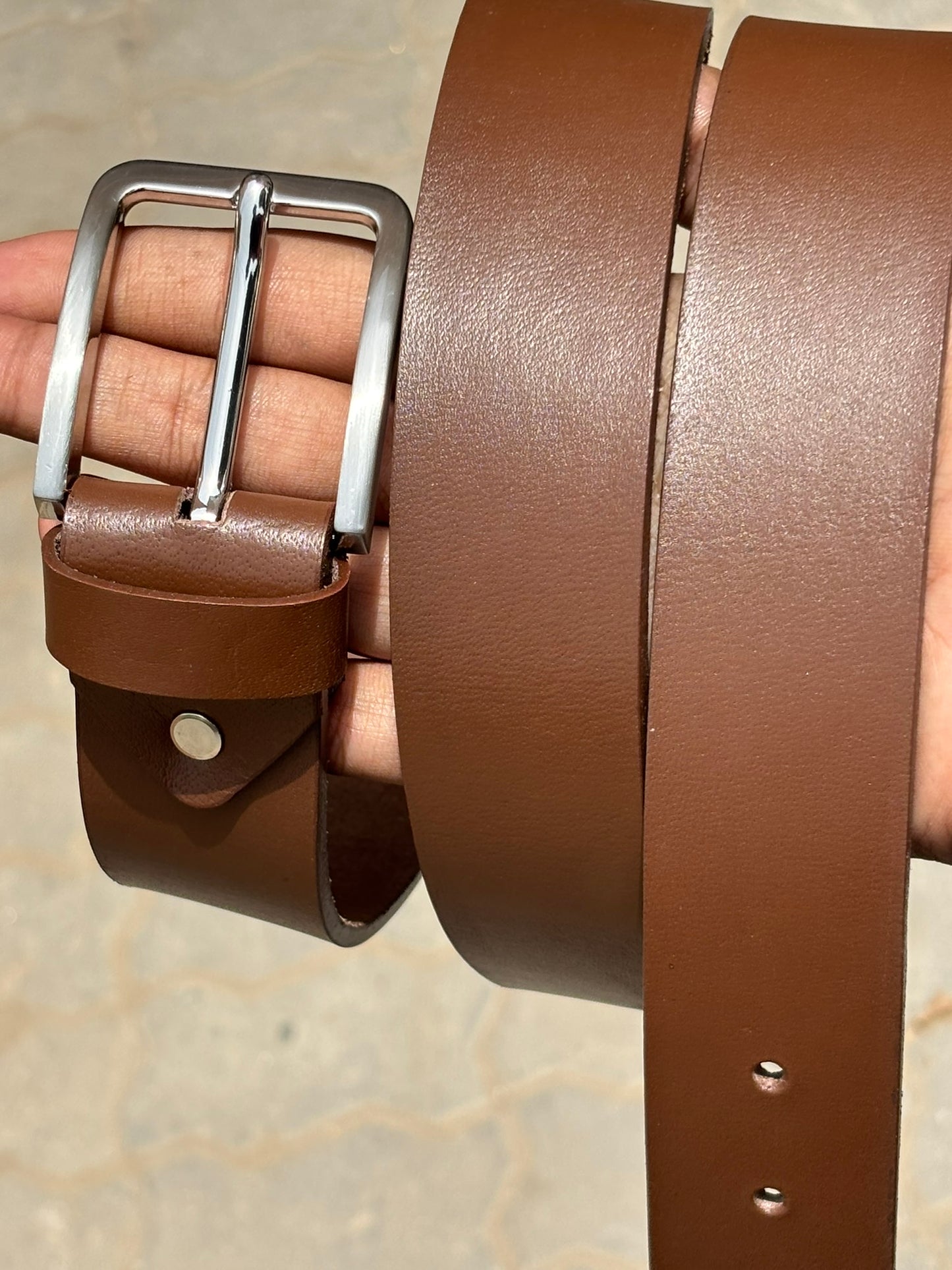 Brown leather belt with a silver buckle held by a hand on a neutral background