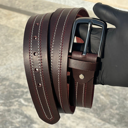 Brown leather belt with a black buckle held by a gloved hand on a gray surface.